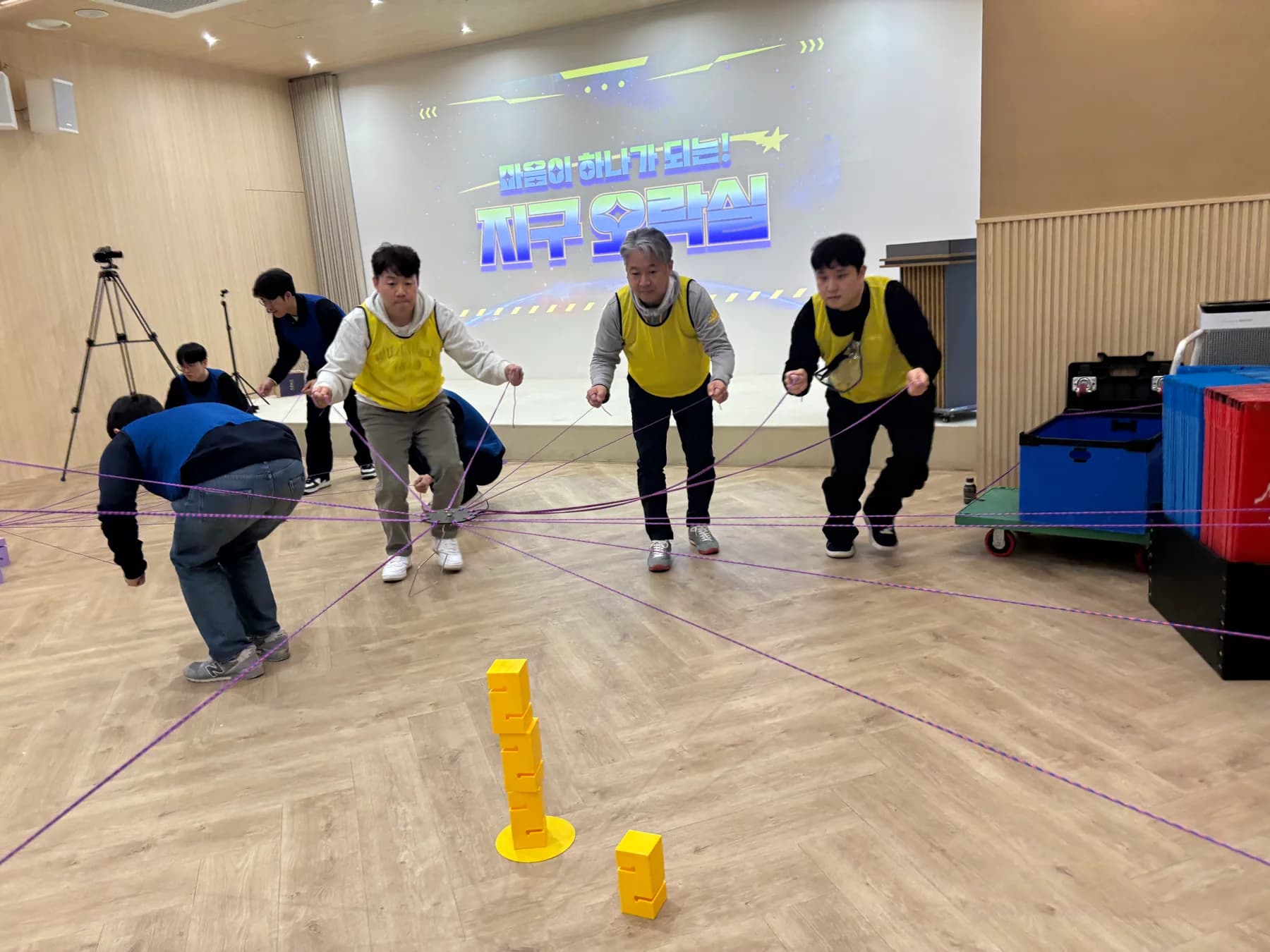 노란 팀 참가자들이 여러 줄을 함께 조작해 블록 탑을 쌓는 실제 협동 경기